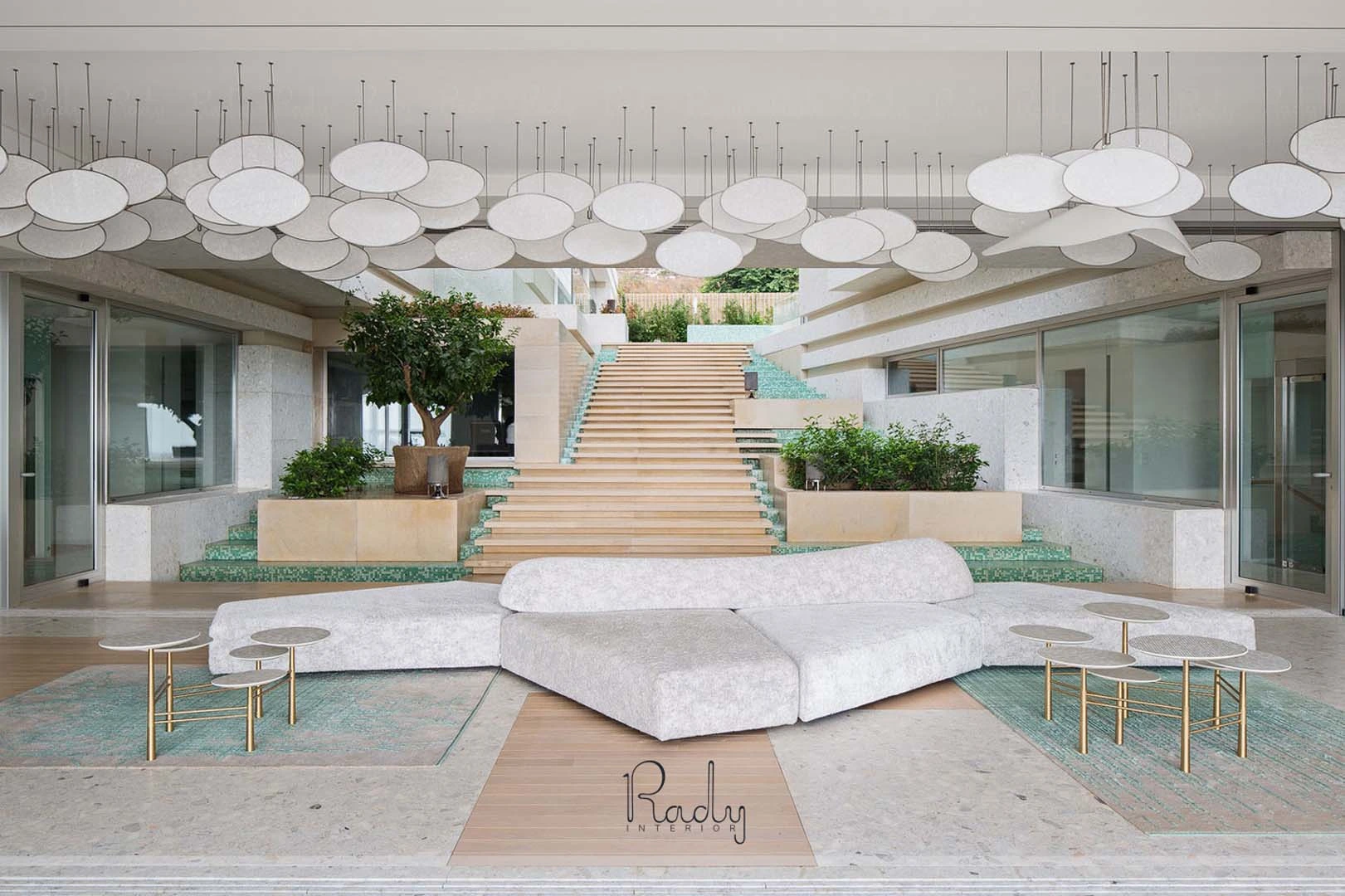 Modern lobby with a white sectional sofa, round tables, two trees, central stairs, and circular ceiling lights. Marble and glass elements create an open, airy design.